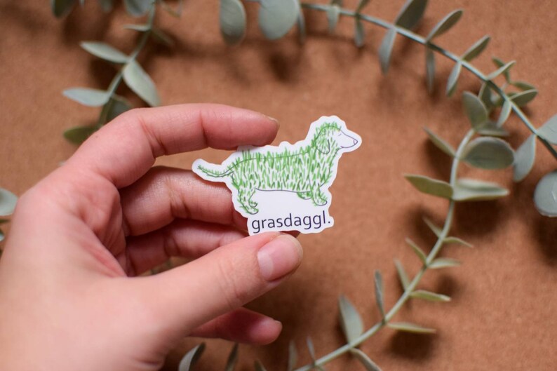 May include: A white sticker with a green line drawing of a dachshund covered in grass. The text "grassdaggl." is below the dog.