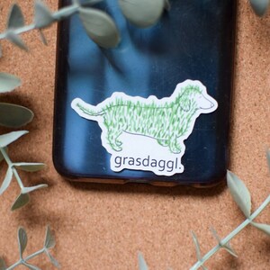 May include: A sticker of a green dachshund with the text "grasdaggl." The sticker is on a black phone case.