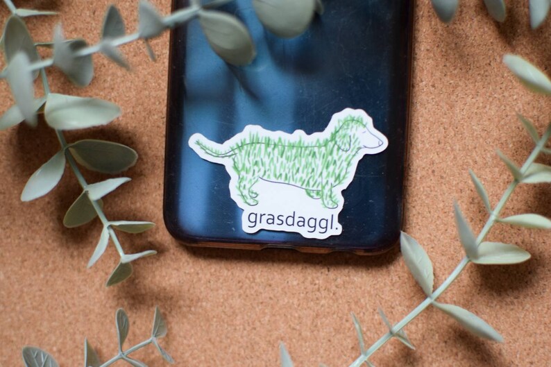 May include: A sticker of a green dachshund with the text "grasdaggl." The sticker is on a black phone case.