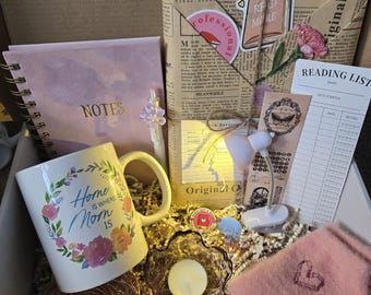 May include: A gift box filled with items for a cozy night in, including a mug with the text "Home is where Mom is", a pink fuzzy blanket, a small lamp, a candle, a notebook, and a reading list.