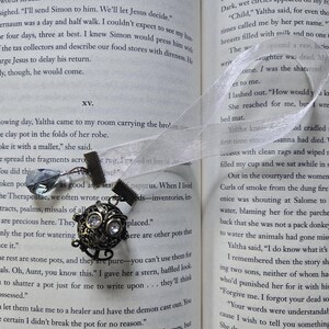 May include: A decorative bookmark with a bronze-colored charm and a clear crystal-like bead. The charm has a detailed, ornate design with clear accents. A sheer white ribbon is attached, resting on the pages of an open book.