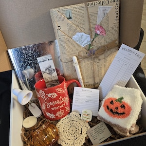 May include: A white gift box filled with various items, including a red mug with the text "Sweater Weather", a small lamp, a candle, a crocheted coaster, a pair of white socks with a pumpkin face, a book review form, and a reading list. The box is filled with brown packing material.