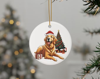 May include: A round, white Christmas ornament featuring a Golden Retriever wearing a Santa hat. The dog is lying down next to a decorated Christmas tree and a wrapped gift. A gold string is attached for hanging.