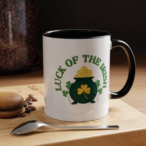 May include: White ceramic mug with a black interior and handle. The mug features a green pot of gold with a shamrock and the text "LUCK OF THE IRISH" in green. A spoon and a macaron are in the foreground.