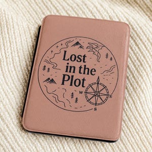 Może przedstawiać: Etui w kolorze przydymionego różu z napisem "Lost in the Plot" w okrągłym wzorze. Projekt zawiera kompas, góry, drzewa i kręte ścieżki. Etui jest prawdopodobnie przeznaczone do czytnika e-booków lub tabletu.