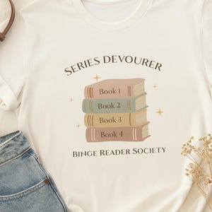 May include: Cream-colored t-shirt with the text "Series Devourer" above a stack of four books labeled "Book 1" through "Book 4." The phrase "Binge Reader Society" is below the books. The shirt is paired with blue jeans.
