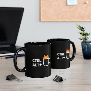 Pode incluir: Duas canecas de cerâmica preta com texto branco e um desenho de gato de desenho animado. As canecas têm o texto "CTRL ALT +" e um gato dentro de um desenho de caneca. As canecas estão em uma superfície de madeira clara.