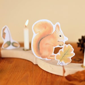 Puede incluir: Un portavelas de madera con un diseño caprichoso que presenta una ardilla, una bellota y una hoja. El soporte tiene tres velas blancas y está colocado sobre una superficie de madera. La ardilla sostiene una hoja.