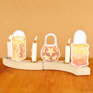 Puede incluir: Un portavelas de madera con tres velas encendidas y tres recortes de farolillos de papel. Los farolillos tienen diseños con temática otoñal, incluyendo hojas y patrones de estrellas. El fondo es de color naranja liso.