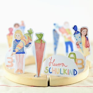 Pode incluir: Um brinquedo educativo de madeira com ilustrações em aquarela de crianças com cones escolares e o texto "Hurra SCHULKIND". As figuras estão dispostas em torno de uma base de madeira, com as letras A, B e o número 3.