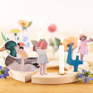 May include: A wooden candle holder with a lit white candle, a blue number 4, and floral accents. Decorative paper cutouts include a duck with children, a girl holding a strawberry, and a girl in a dress. The candle holder is on a wooden surface.