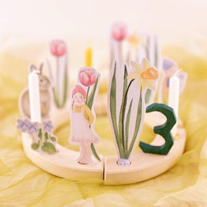 May include: A wooden cake topper with a spring theme. It features a girl, flowers, a bunny, and the number 3. The topper is perfect for a birthday cake.