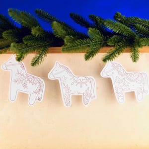 May include: A garland of three white horse decorations with pink floral patterns. The ornaments are strung together, set against a blue backdrop. Green fir branches are at the top.