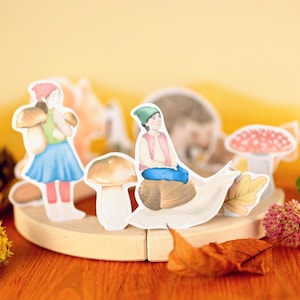 Puede incluir: Una exhibición de madera caprichosa con recortes de papel de personajes con temática de bosque. Una niña con falda azul lleva setas, y otra monta un caracol. Otros recortes incluyen una seta, un erizo y hojas de otoño. El fondo es amarillo cálido.