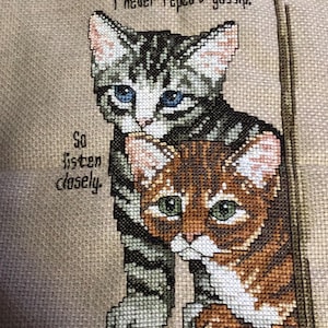 May include: Cross-stitch artwork featuring two cats. One cat is gray and black striped with blue eyes, and the other is brown and orange with green eyes. Text reads, "I never repeat gossip. So listen closely."