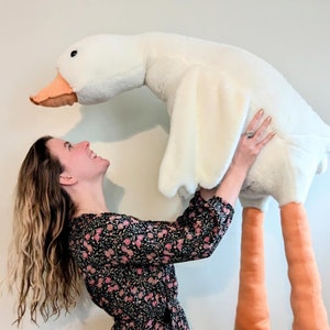 K&ouml;nnte beinhalten: Eine Frau in einem schwarzen Blumenkleid h&auml;lt eine gro&szlig;e wei&szlig;e Pl&uuml;schgans mit orangenen Beinen. Die Gans schaut die Frau an.
