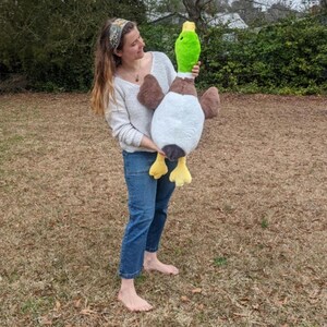 Puede incluir: Un juguete de peluche grande con forma de pato azul&oacute;n que presenta una cabeza verde brillante, pecho marr&oacute;n, vientre blanco y alas de color marr&oacute;n oscuro. El peluche tiene un pico amarillo y patas palmeadas amarillas de tela suave.
