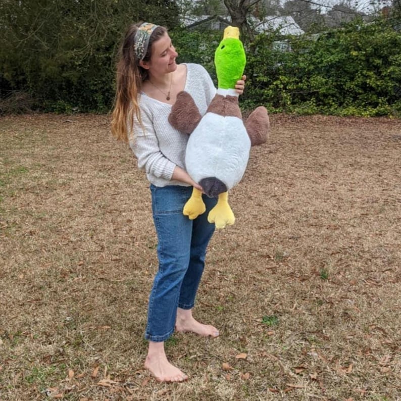 Puede incluir: Un juguete de peluche grande con forma de pato azul&oacute;n que presenta una cabeza verde brillante, pecho marr&oacute;n, vientre blanco y alas de color marr&oacute;n oscuro. El peluche tiene un pico amarillo y patas palmeadas amarillas de tela suave.