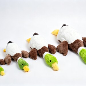 Puede incluir: Cuatro juguetes de peluche con forma de pato real, dispuestos en fila del m&aacute;s peque&ntilde;o al m&aacute;s grande. Cada pato tiene un cuerpo blanco, alas marrones, una cabeza de color verde brillante y un pico amarillo. Los peluches est&aacute;n sobre un fondo blanco.
