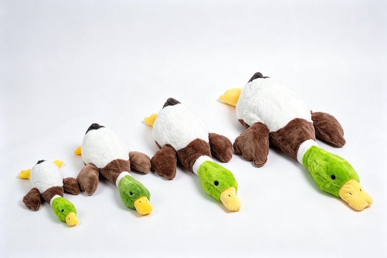 Puede incluir: Cuatro juguetes de peluche con forma de pato real, dispuestos en fila del m&aacute;s peque&ntilde;o al m&aacute;s grande. Cada pato tiene un cuerpo blanco, alas marrones, una cabeza de color verde brillante y un pico amarillo. Los peluches est&aacute;n sobre un fondo blanco.