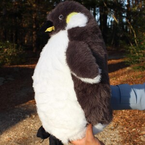 May include: A plush penguin toy featuring soft dark brown and white fur, a black beak with a yellow accent, and black feet. The stuffed animal has a rounded body, a white chest, and a dark brown head with a small yellow patch near the eye area.