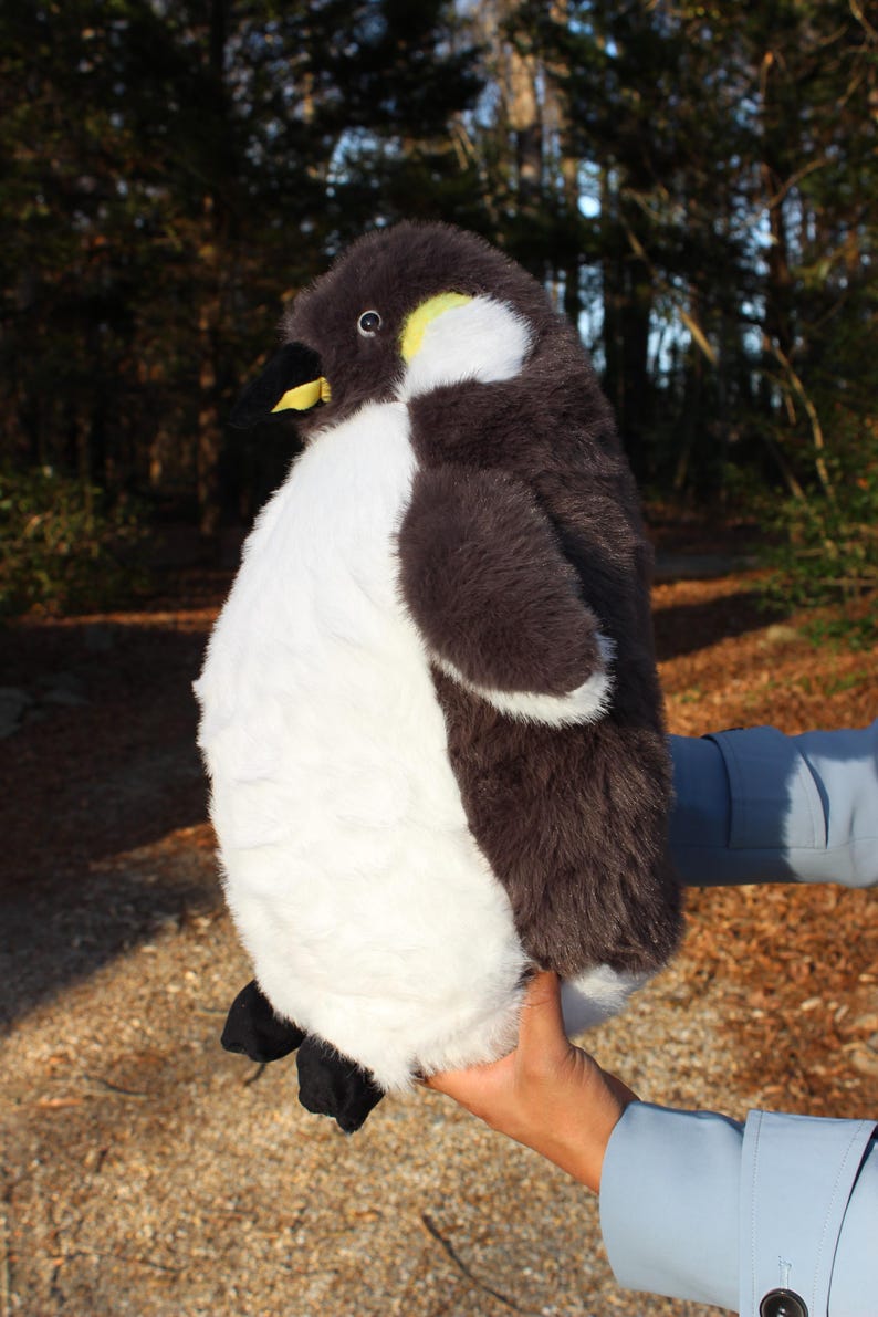 May include: A plush penguin toy featuring soft dark brown and white fur, a black beak with a yellow accent, and black feet. The stuffed animal has a rounded body, a white chest, and a dark brown head with a small yellow patch near the eye area.