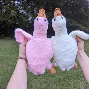 K&ouml;nnte beinhalten: Zwei Pl&uuml;sch-G&auml;nse-Spielzeuge, eines rosa und eines wei&szlig;, werden in die Luft gehalten. Die G&auml;nse haben braune Schn&auml;bel und flauschige wei&szlig;e Federn.