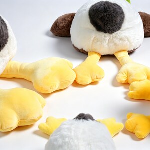 Puede incluir: Una colecci&oacute;n de juguetes de peluche con forma de pato que presentan cuerpos blancos, manchas de color gris oscuro en la cabeza, alas marrones y patas palmeadas de color amarillo brillante. Los peluches est&aacute;n dispuestos en grupo.