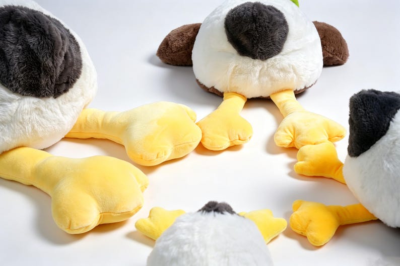 Puede incluir: Una colecci&oacute;n de juguetes de peluche con forma de pato que presentan cuerpos blancos, manchas de color gris oscuro en la cabeza, alas marrones y patas palmeadas de color amarillo brillante. Los peluches est&aacute;n dispuestos en grupo.