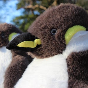 May include: Two plush penguin toys featuring soft dark brown fur on the head and back, white fur on the chest, and black beaks with bright yellow accents. Each penguin has a small dark eye and a rounded, fuzzy texture throughout the body.