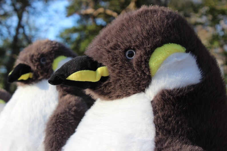 May include: Two plush penguin toys featuring soft dark brown fur on the head and back, white fur on the chest, and black beaks with bright yellow accents. Each penguin has a small dark eye and a rounded, fuzzy texture throughout the body.