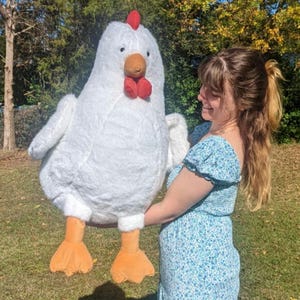 May include: A large, plush white chicken stuffed animal with a soft, fuzzy texture. It features a bright red comb on top of its head, a brown beak, and two dangling red wattles. The chicken has two orange, webbed feet extending from the bottom.