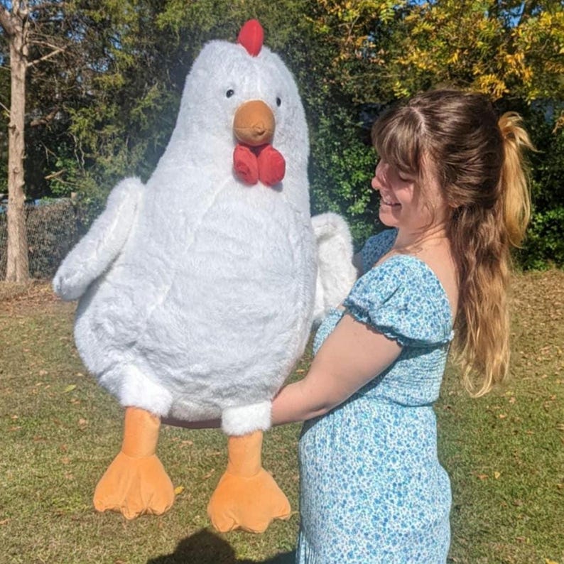 May include: A large, plush white chicken stuffed animal with a soft, fuzzy texture. It features a bright red comb on top of its head, a brown beak, and two dangling red wattles. The chicken has two orange, webbed feet extending from the bottom.