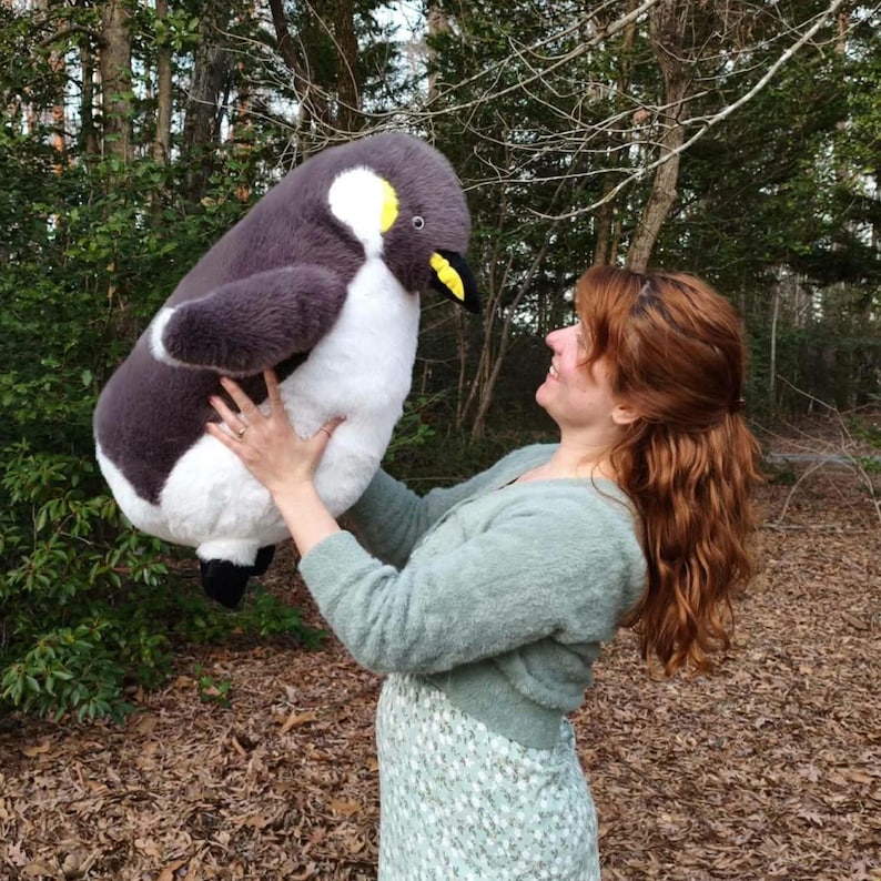 May include: A large plush penguin toy featuring a soft, dark gray back, a white belly, a yellow patch near the eye, and a black beak. The stuffed animal has a rounded, plump body shape and a textured, fur-like exterior finish throughout.