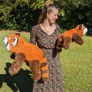 May include: A woman is holding two plush red panda toys. The toys are brown and orange with black stripes on their tails. The woman is wearing a floral dress.