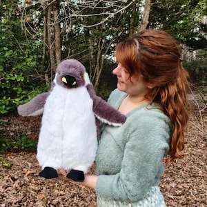 May include: A plush penguin toy featuring a soft white belly, dark gray wings, and a dark gray head with yellow accents near the eyes. The stuffed animal has a black beak and small black feet, designed with a fluffy texture throughout its body.