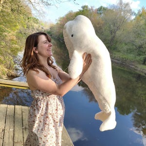 May include: A large, plush white beluga whale stuffed animal. The toy has a rounded head, flipper-like appendages, and a tail. The plushie is being held by a person in a floral dress near a body of water.