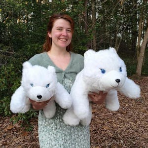 May include: Two white plush polar bear stuffed animals with blue eyes and black noses are held up in front of a person. The bears have a fluffy texture and are positioned to face forward. The background is a natural outdoor setting with trees.