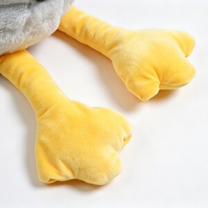 Puede incluir: Un juguete de peluche con un cuerpo superior gris de textura suave y dos piernas largas de color amarillo brillante con pies anchos y palmeados. La tela tiene una superficie difusa y t&aacute;ctil, y las extremidades amarillas son lisas.