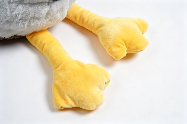 Puede incluir: Un juguete de peluche con un cuerpo superior gris de textura suave y dos piernas largas de color amarillo brillante con pies anchos y palmeados. La tela tiene una superficie difusa y t&aacute;ctil, y las extremidades amarillas son lisas.