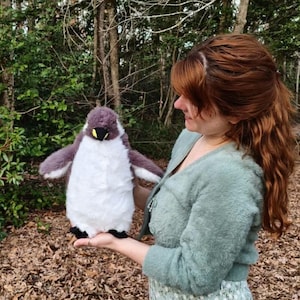 May include: A plush penguin toy featuring a soft, dark gray back and wings with a contrasting white chest and belly. The stuffed animal has a small, pointed black beak with a yellow accent at the base and black feet, designed with a fuzzy texture.