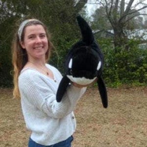 May include: A plush orca whale toy in black and white. The toy has a rounded body, a dorsal fin, and two side fins. The orca's face has white markings around the eyes and mouth. The toy is held up in front of a person.