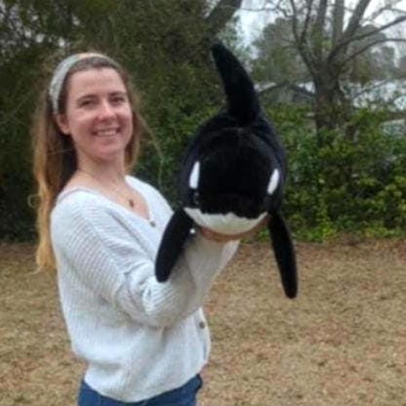 May include: A plush orca whale toy in black and white. The toy has a rounded body, a dorsal fin, and two side fins. The orca's face has white markings around the eyes and mouth. The toy is held up in front of a person.