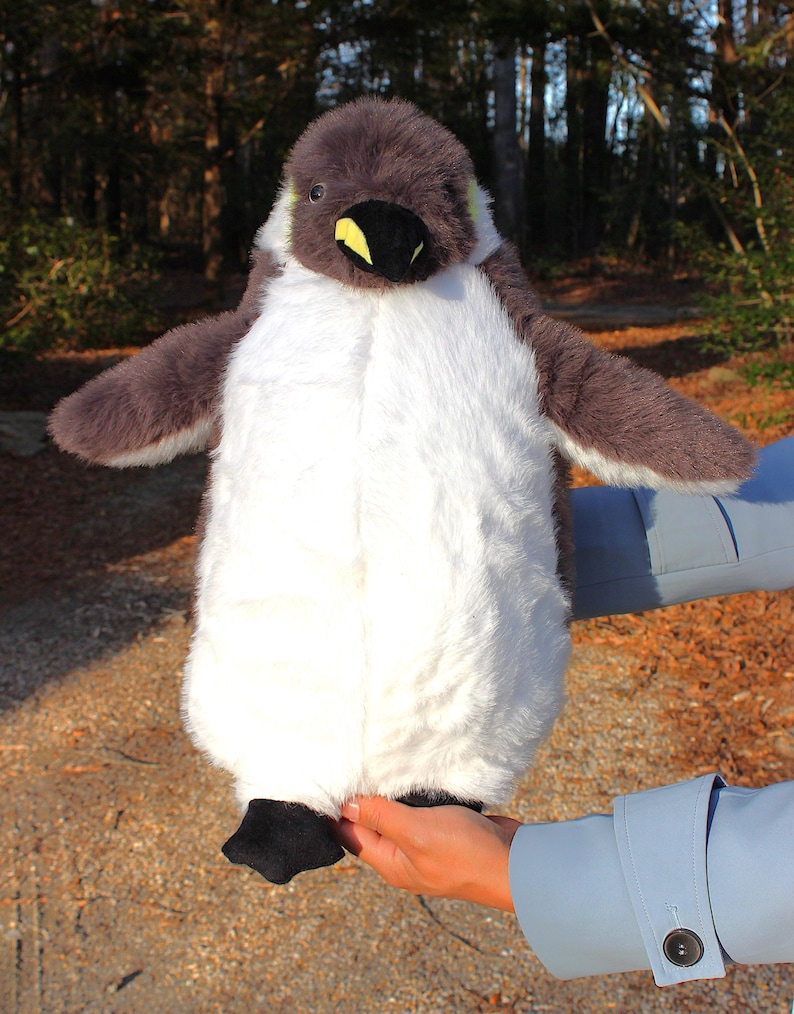 May include: A plush penguin toy featuring soft, fluffy white fur on the chest and belly, contrasted by dark gray fur on the head, back, and wings. The penguin has a black beak with a small yellow accent, dark eyes, and black fabric feet.