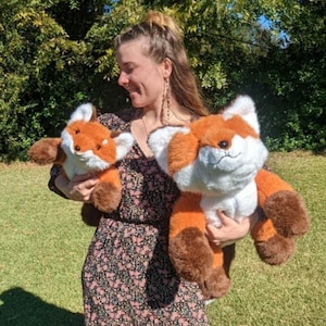 May include: A woman is holding two plush fox toys. The larger fox is brown and orange with white fur on its face. The smaller fox is orange with white fur on its face. Both foxes have black eyes and black noses.