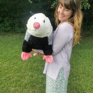 May include: A woman is holding a large plush opossum toy. The opossum is black, white, and gray with pink feet and a pink nose. The woman is wearing a light purple shirt and a green and white floral skirt.
