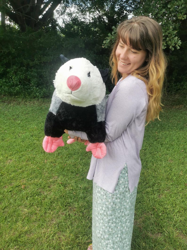 May include: A woman is holding a large plush opossum toy. The opossum is black, white, and gray with pink feet and a pink nose. The woman is wearing a light purple shirt and a green and white floral skirt.
