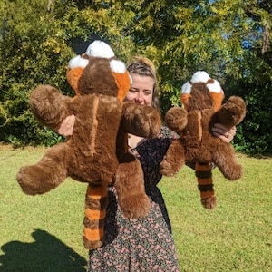 May include: Two plush red panda toys, one large and one small, are being held by a person. The larger plush toy is brown with orange and white markings. The smaller plush toy is brown with orange and white markings. Both plush toys have long tails.
