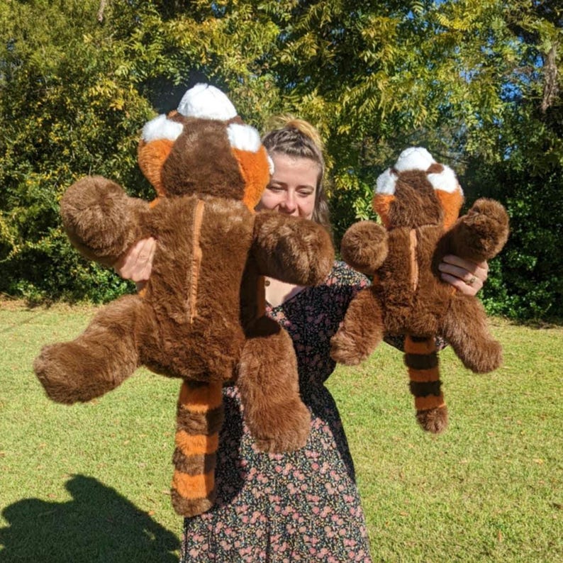May include: Two plush red panda toys, one large and one small, are being held by a person. The larger plush toy is brown with orange and white markings. The smaller plush toy is brown with orange and white markings. Both plush toys have long tails.
