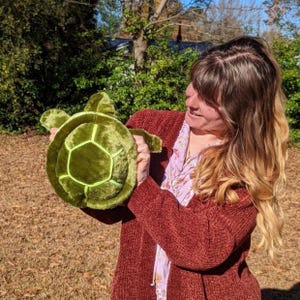 May include: A woman is holding a green plush turtle toy. The turtle has a light green shell with a textured surface.